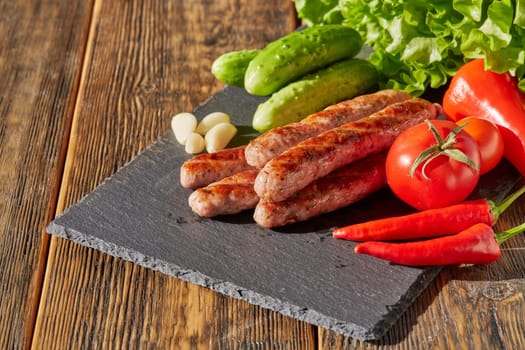 Grilled pork sausages  with fresh vegetables  on stone cutting board on wooden background.