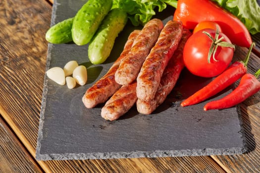 Grilled pork sausages  with fresh vegetables  on stone cutting board on wooden background.
