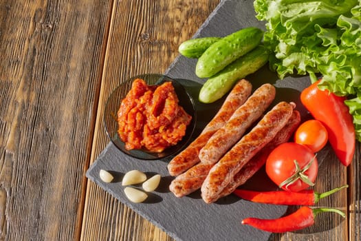 Grilled pork sausages  with fresh vegetables  on stone cutting board on wooden background. Top view