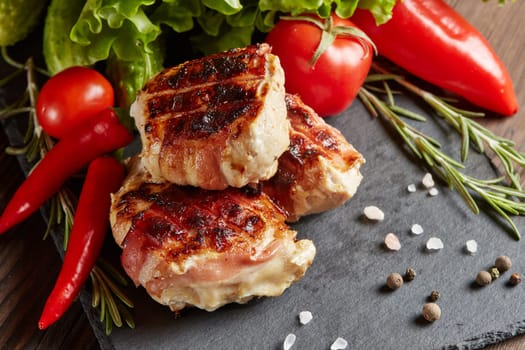 Bacon-wrapped grilled chicken (turkey)  fillet with fresh vegetables and spices on black stone cutting board on