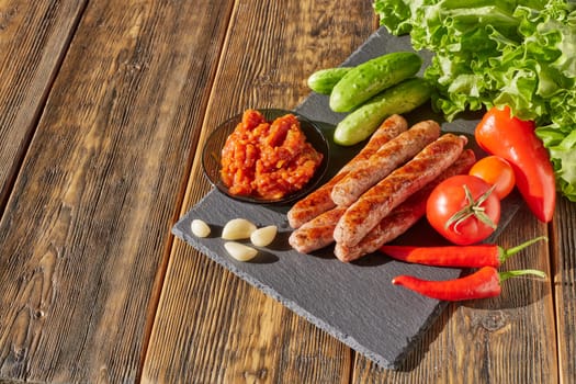 Grilled pork sausages  with fresh vegetables  on stone cutting board on wooden background. Top view with copy space.