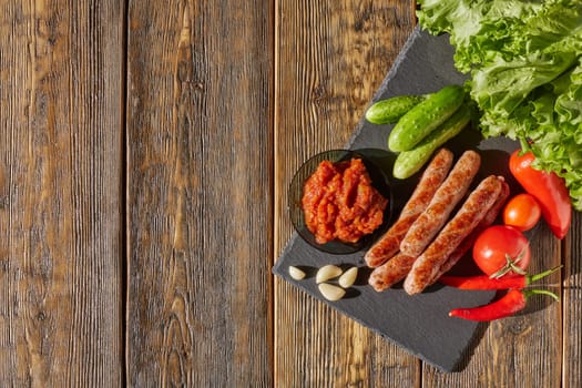Grilled pork sausages  with fresh vegetables  on stone cutting board on wooden background. Top view with copy space.