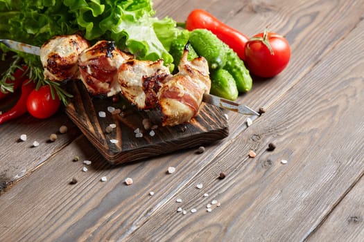 Shashlik from chicken with bacon. Grilled chicken (turkey) fillet on skewer and fresh vegetables with spices on stone cutting board on wooden background