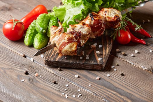 Shashlik from chicken with bacon. Grilled chicken (turkey) fillet on skewer and fresh vegetables with spices on stone cutting board on wooden background