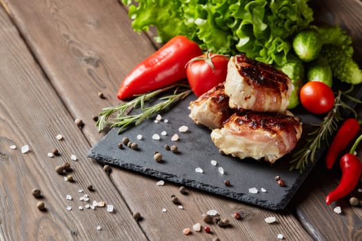 Bacon-wrapped grilled chicken (turkey) fillet with fresh vegetables and spices on stone cutting board on wooden background