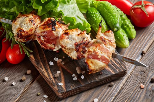 Shashlik from chicken with bacon. Grilled chicken (turkey) fillet on skewer and fresh vegetables with spices on stone cutting board on wooden background