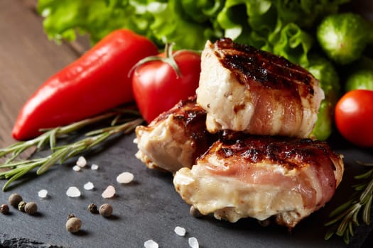 Bacon-wrapped grilled chicken (turkey) fillet with fresh vegetables and spices on stone cutting board on wooden background