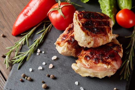 Bacon-wrapped grilled chicken (turkey) fillet with fresh vegetables and spices on stone cutting board on wooden background