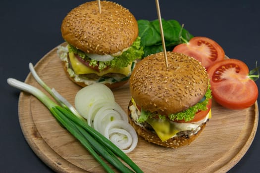 Two hamburgers close-up on a round wooden board, next to, tomatoes, chopped onions and spinach leaves.