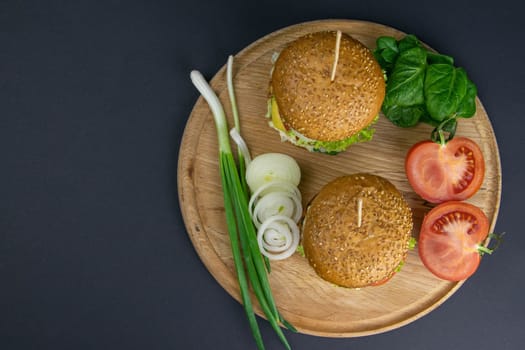 Two hamburgers close-up on a round wooden board, next to, tomatoes, chopped onions and spinach leaves. Top views with clear space