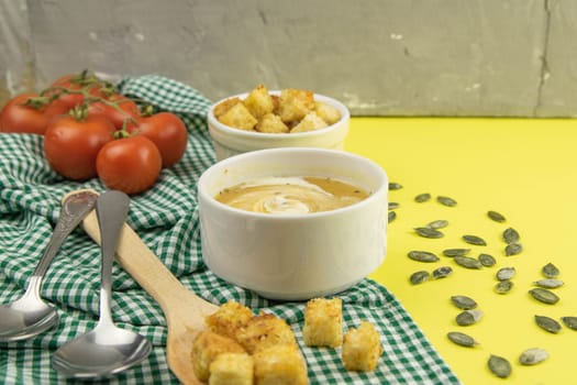 Pea soup with croutons stands on a green towel, next to there are tomatoes, toasts of pumpkin seeds, everything stands on the yellow table.