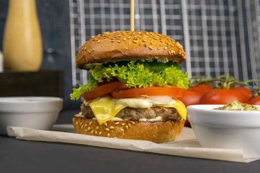 Close-up hamburger, sauces, french fries and tomatoes stand nearby, grill rack in the background.