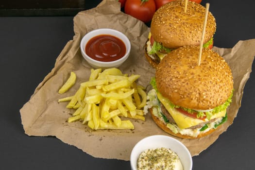 Close-up hamburger, sauces, french fries and tomatoes stand nearby, grill rack in the background.