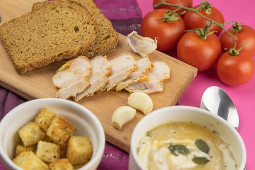 Pea soup with toasts and pumpkin seeds, next to the wooden board sliced bacon, garlic and black bread, next to tomatoes.
