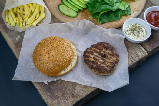 Preparing for the preparation of hamburgers. On a wooden board are lettuce leaves, chopped: tomatoes, cucumbers, onions, cutlets and sauces.