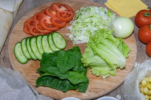 Preparing for the preparation of hamburgers. On a wooden board are lettuce leaves, chopped: tomatoes, cucumbers, onions, cutlets and sauces.
