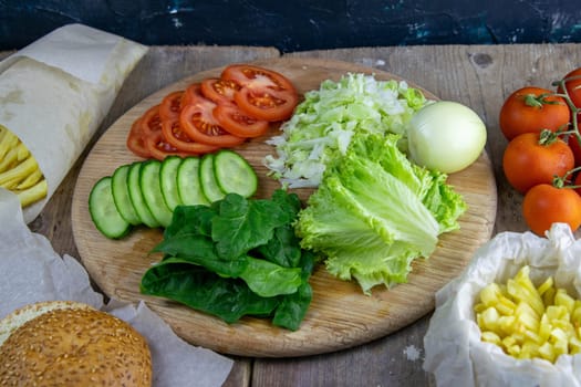 Preparing for the preparation of hamburgers. On a wooden board are lettuce leaves, chopped: tomatoes, cucumbers, onions, cutlets and sauces.