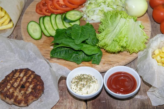 Preparing for the preparation of hamburgers. On a wooden board are lettuce leaves, chopped: tomatoes, cucumbers, onions, cutlets and sauces.