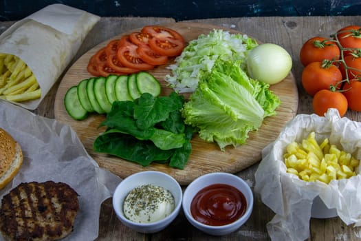Preparing for the preparation of hamburgers. On a wooden board are lettuce leaves, chopped: tomatoes, cucumbers, onions, cutlets and sauces.