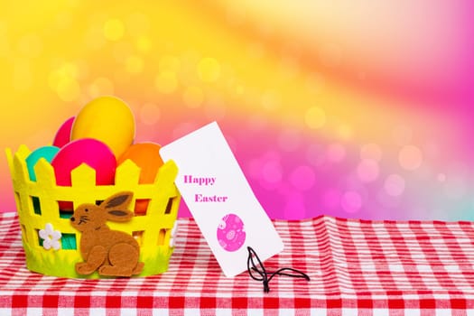 Table top on easter background. Colorful easter eggs in a basket with a bunny and a easter tag on picnic tablecloth over abstract rainbow backdrop. For your food and product display montage. Space.