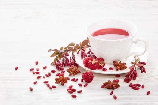 Healthy fruit tea with anise, barberries and raspberries in white porcelain cup and branch of berberry on light wooden background.
