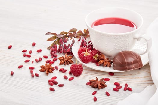 Healthy fruit tea with anise, barberries and raspberries in white porcelain cup and branch of berberry on light wooden background.