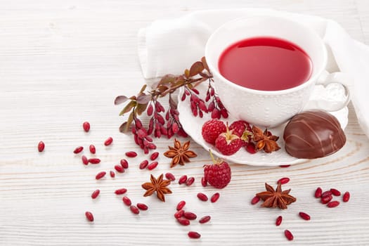 Healthy fruit tea with anise, barberries and raspberries in white porcelain cup and branch of berberry on light wooden background.