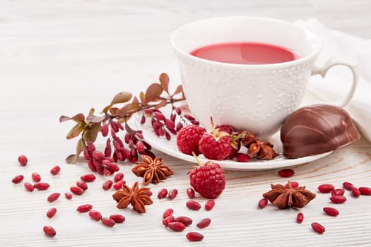 Healthy fruit tea with anise, barberries and raspberries in white porcelain cup and branch of berberry on light wooden background.