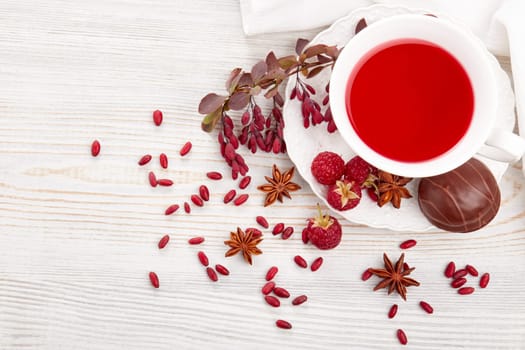 Healthy fruit tea with anise, barberries and raspberries in white porcelain cup and branch of berberry on light wooden background. Top view