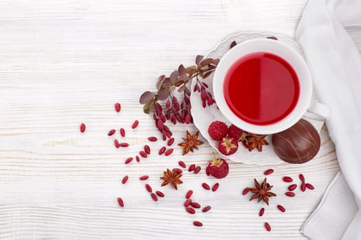 Healthy fruit tea with anise, barberries and raspberries in white porcelain cup and branch of berberry on light wooden background. Top view