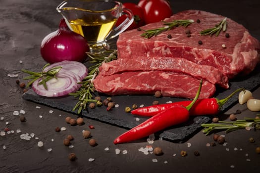 Still life with raw meat beef fillet on black stone cutting board, rosemary twigs, set of different peppers, vegatables and olive oil on dark background. Fresh beef piece for steak or grilled barbecue