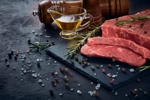 Still life with raw meat beef fillet on black stone cutting board,  rosemary twigs, set of different peppers, salt and olive oil on dark background. Fresh beef piece for steak or grilled barbecue