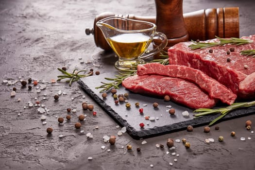 Still life with raw meat beef fillet on black stone cutting board,  rosemary twigs, set of different peppers, salt and olive oil on dark background. Fresh beef piece for steak or grilled barbecue