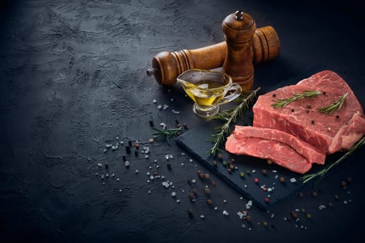 Raw meat beef fillet on black stone cutting board,  rosemary, set of different peppers, salt and olive oil on dark background. Fresh beef piece for steak or grilled barbecue. Top view with copy space