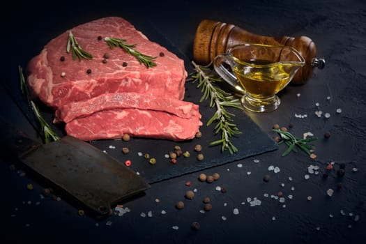 Still life with raw meat beef fillet on black stone cutting board,  rosemary twigs, set of different peppers, salt and olive oil on dark background. Fresh beef piece for steak or grilled barbecue