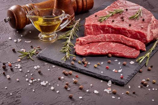 Still life with raw meat beef fillet on black stone cutting board,  rosemary twigs, set of different peppers, salt and olive oil on dark background. Fresh beef piece for steak or grilled barbecue