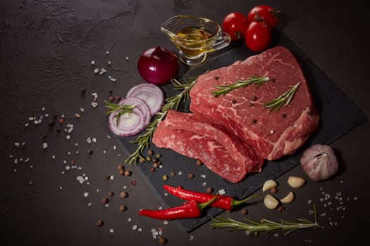 Raw meat beef fillet on stone cutting board, rosemary, set of different peppers, vegatables, olive oil on dark background. Fresh beef piece for steak or grilled barbecue. Top view with copy space