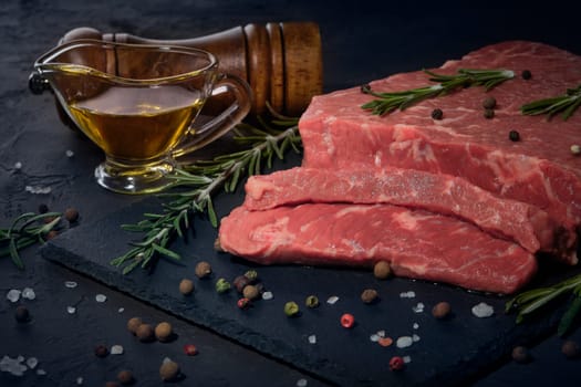 Still life with raw meat beef fillet on black stone cutting board,  rosemary twigs, set of different peppers, salt and olive oil on dark background. Fresh beef piece for steak or grilled barbecue