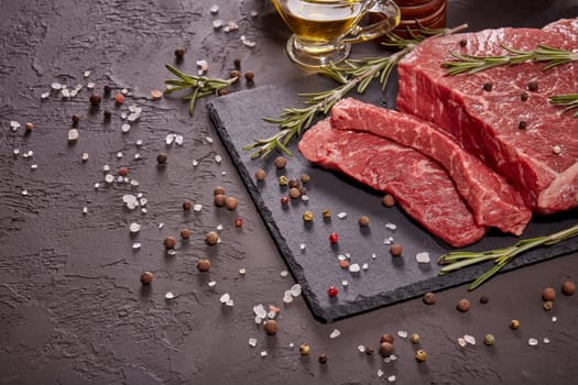 Raw meat beef fillet on black stone cutting board,  rosemary, set of different peppers, salt and olive oil on dark background. Fresh beef piece for steak or grilled barbecue. Top view with copy space