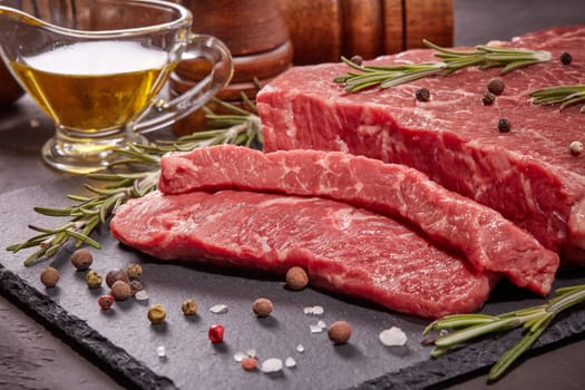 Still life with raw meat beef fillet on black stone cutting board,  rosemary twigs, set of different peppers, salt and olive oil on dark background. Fresh beef piece for steak or grilled barbecue