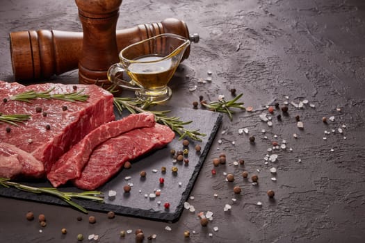 Raw meat beef fillet on black stone cutting board,  rosemary, set of different peppers, salt and olive oil on dark background. Fresh beef piece for steak or grilled barbecue. Top view with copy space