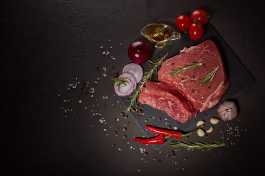Raw meat beef fillet on stone cutting board, rosemary, set of different peppers, vegatables, olive oil on dark background. Fresh beef piece for steak or grilled barbecue. Top view with copy space