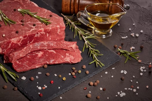 Still life with raw meat beef fillet on black stone cutting board,  rosemary twigs, set of different peppers, salt and olive oil on dark background. Fresh beef piece for steak or grilled barbecue