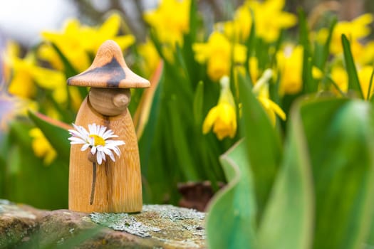 Wooden elf figure with a daisy in hand is standing in a flower bed in front of blurred yellow flowers