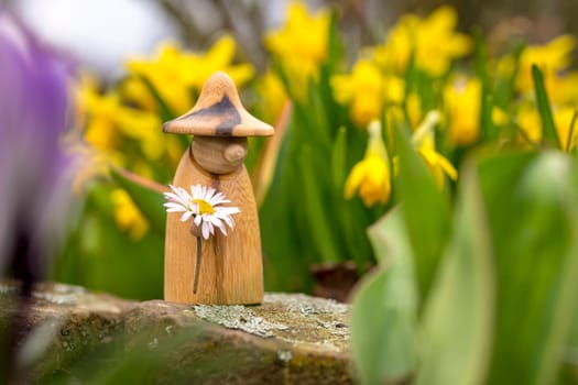 Wooden elf figure with a daisy in hand is standing in a flower bed in front of blurred yellow flowers