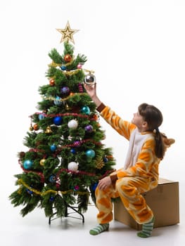 The girl sits on a box in front of the Christmas tree and hangs a Christmas tree ball