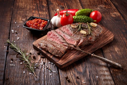 Grilled meat. Medium rare beef steak on wooden cutting board, rosemary twigs, spices and vegatables on brown wooden background. Steak menu