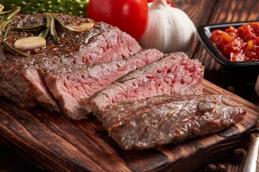 Grilled meat. Medium rare beef steak on wooden cutting board, rosemary twigs, spices and vegatables on brown wooden background. Steak menu