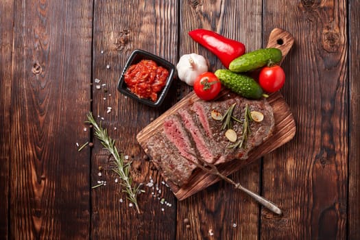 Grilled meat. Medium rare beef steak on wooden cutting board, rosemary twigs, spices and vegatables on brown wooden background. Top view. Steak menu