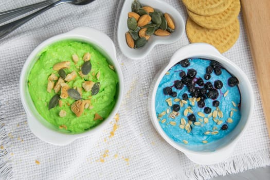 Two curd desserts: one with blueberries and nuts and the other with avocado and kiwi, next to there are pumpkin seeds and nuts.